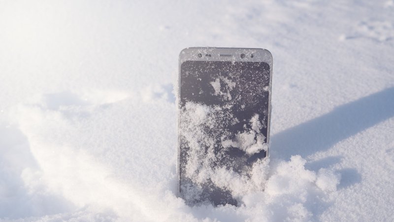 A phone standing upright in the snow, particles of snow covering the screen.